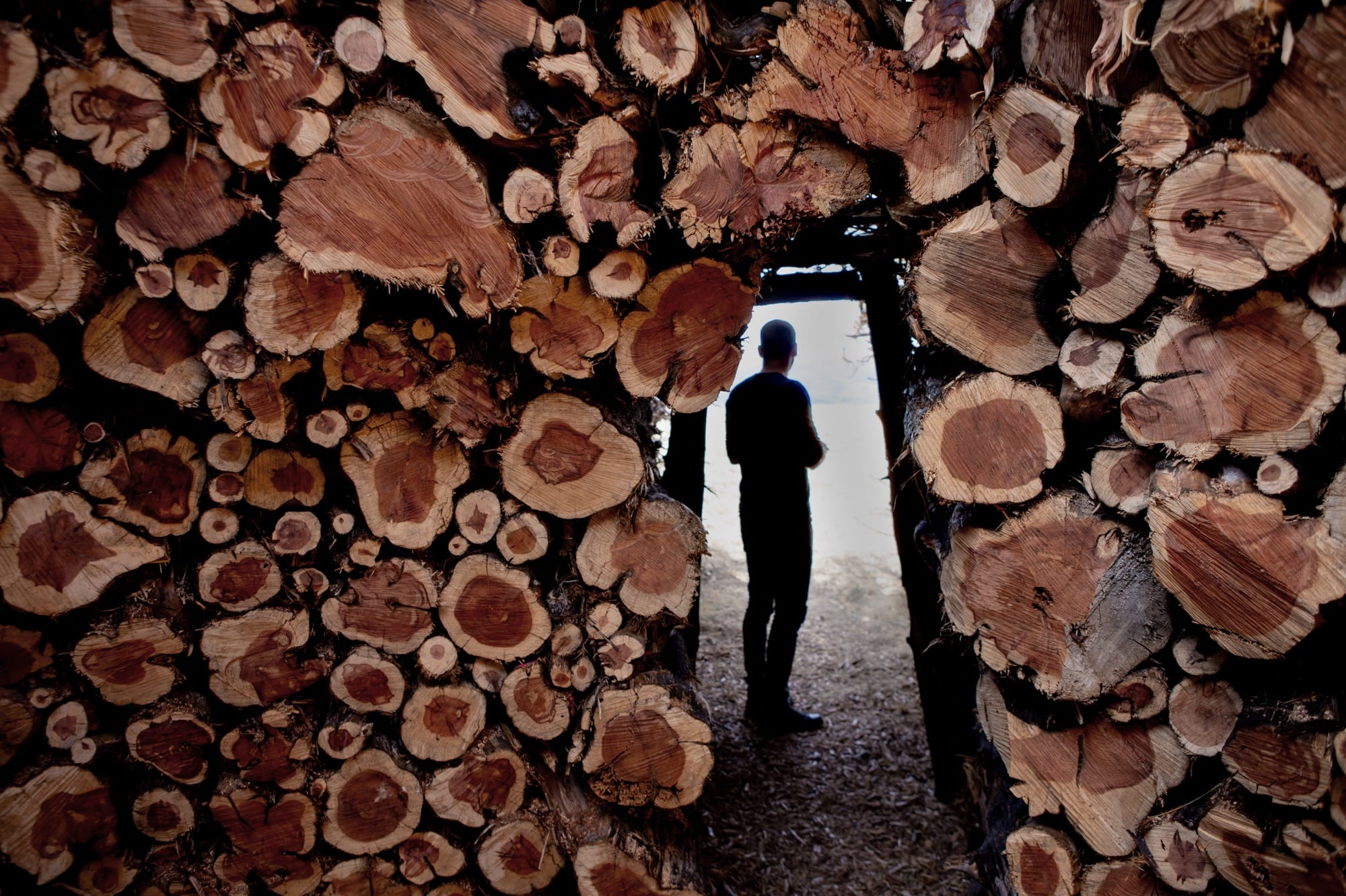 stacked logs form an opening in a structure by nomad studio. a man stands in the opening