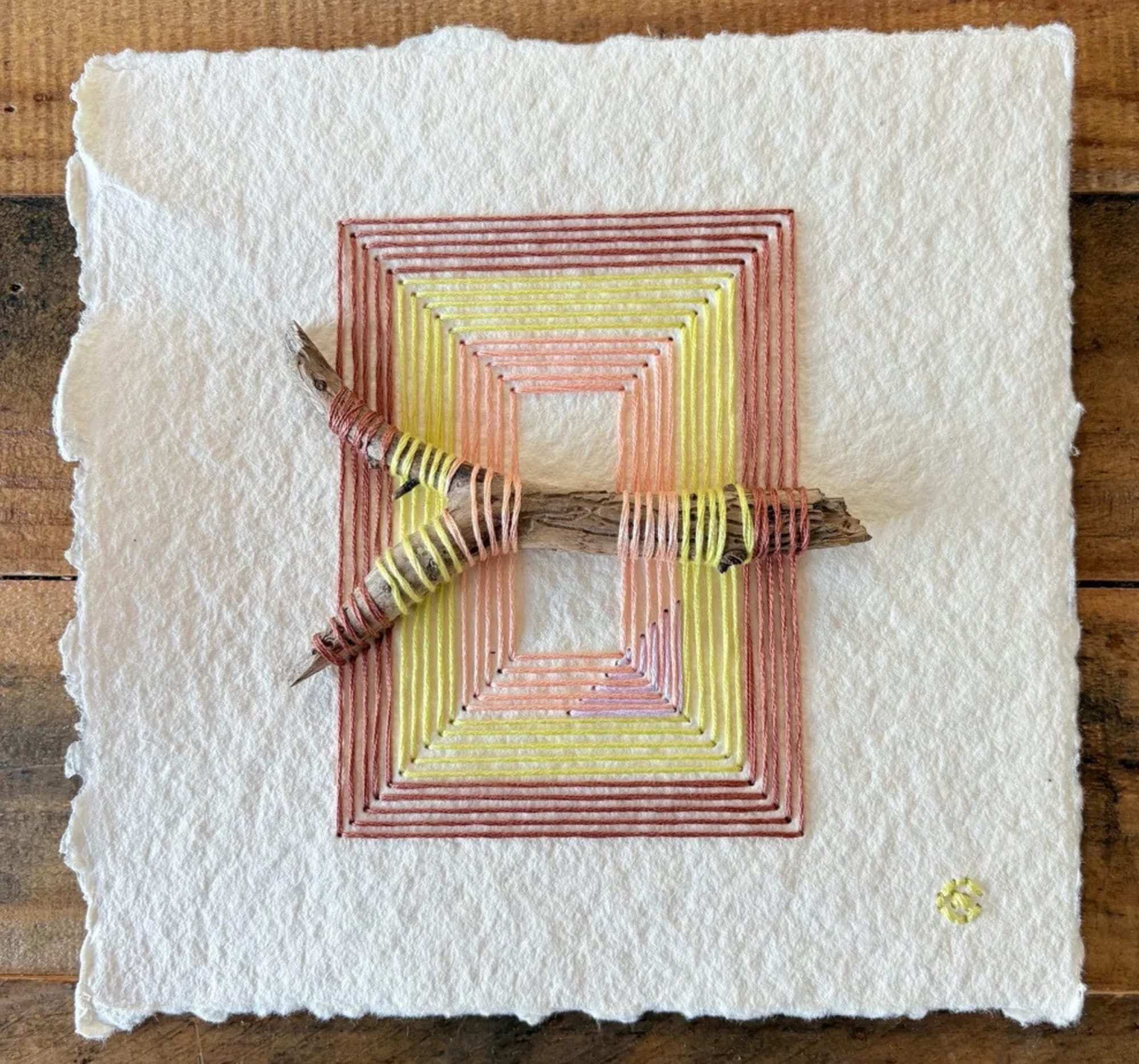 a paper artwork by Natalie Ciccoricco featuring handmade paper and pink, yellow, and ochre thread wound around found twigs in a geometric shape